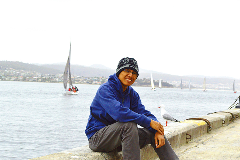 A man wearing black cap and blue jacket is sitting by the bay