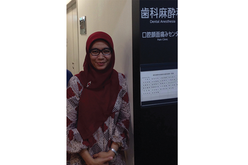A woman with glasses and red hijab wearing batik dress is standing beside Dental Anesthesia signage