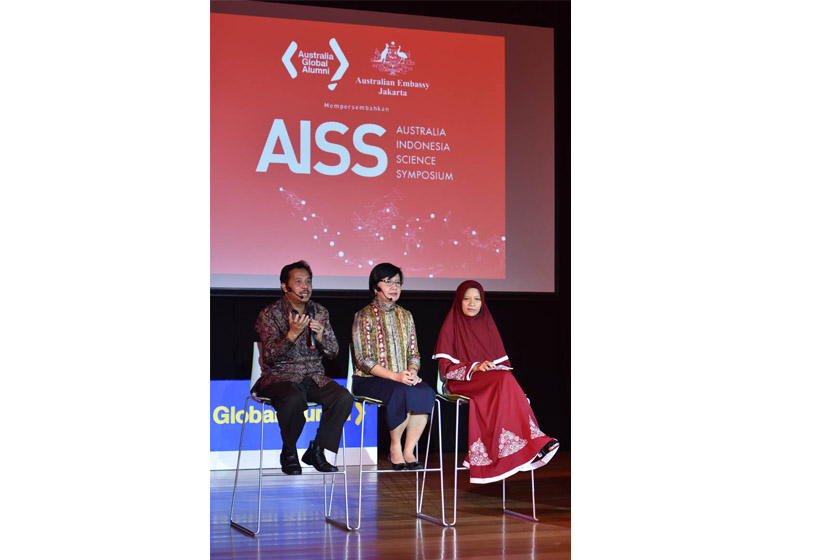 Three people are sitting on a chair on the stage with AISS logo appearing on the screen behind them